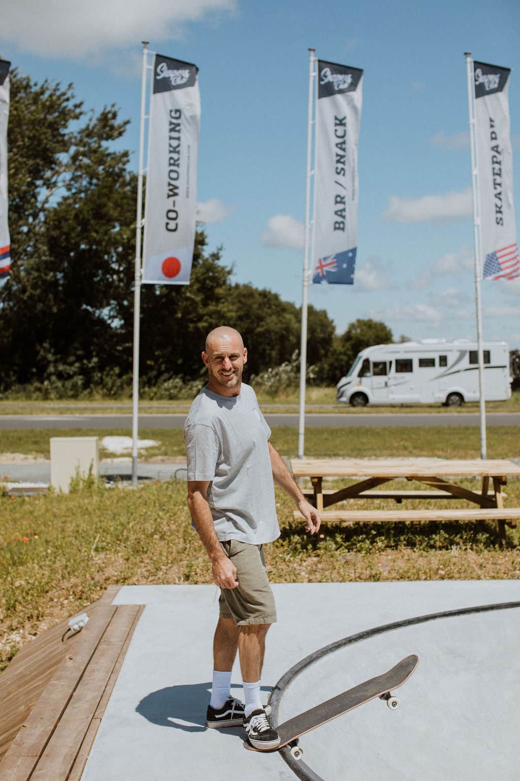 A man in a gray T-shirt and shorts stands with a skateboard at the edge of an outdoor skatepark bowl. Flags, a lawn and a white van are visible in the background under a blue sky.