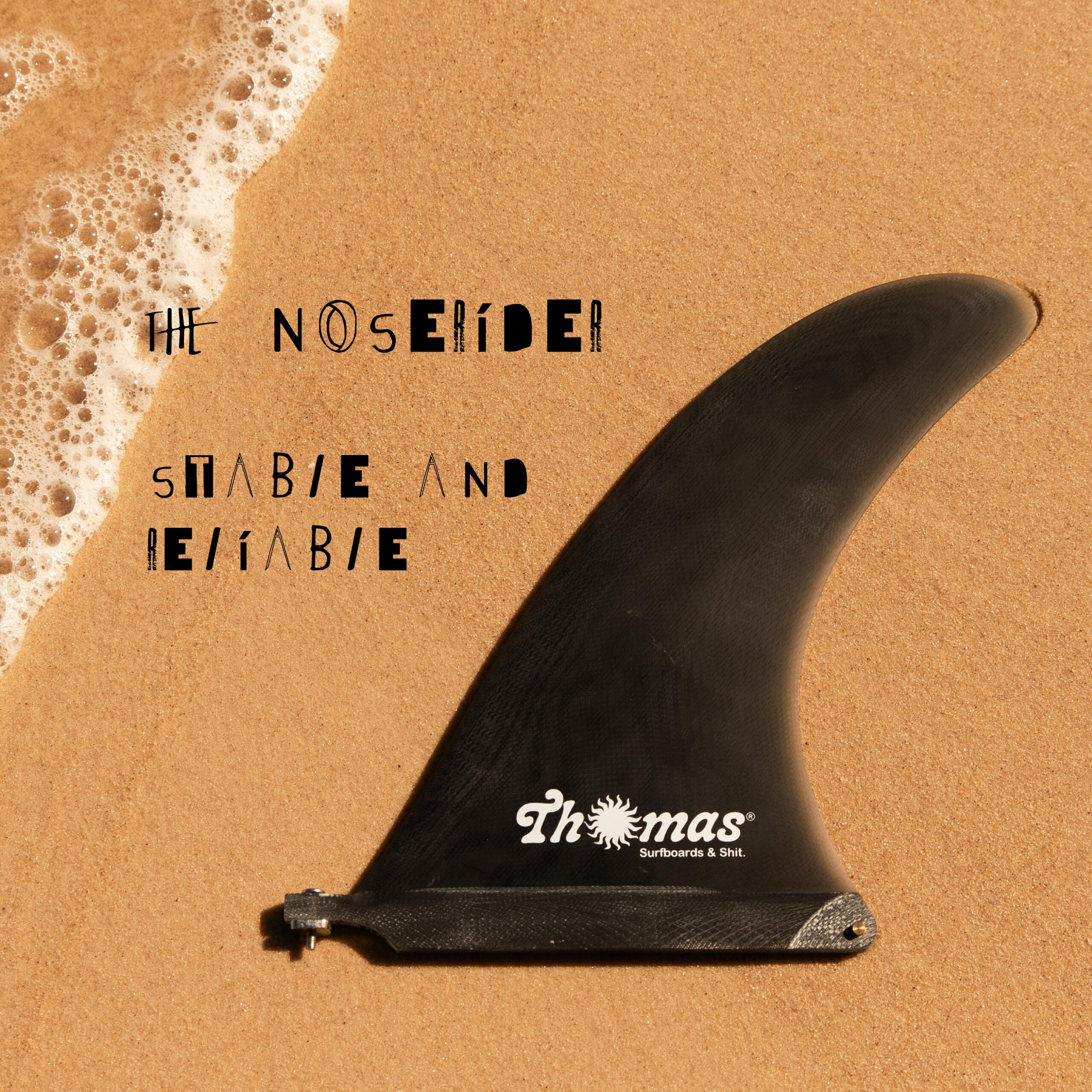Un aileron de planche de surf noir, étiqueté « Thomas Surfboards & Sht. », repose sur le sable mouillé, au bord de l'eau écumeuse de l'océan. À côté, on peut lire : « Le Noserider. Stable et fiable. ».
